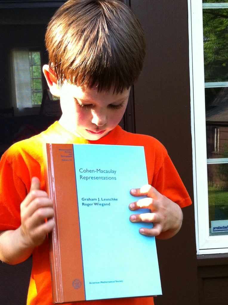 A small boy in an orange shirt holding a copy of Cohen-Macaulay Representations